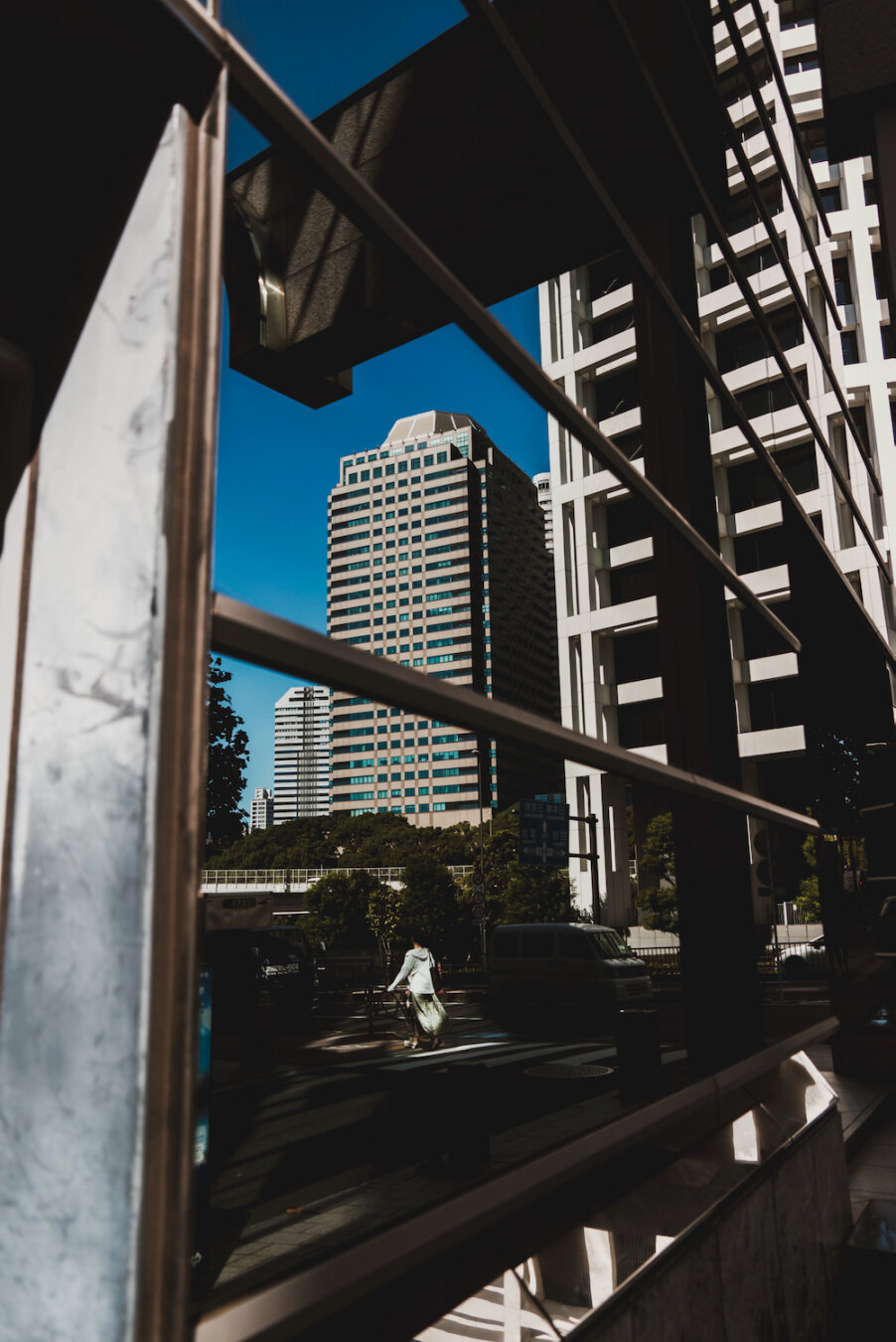 A sharp vertical view capturing the morning light across Moto-Akasaka and Kioicho. High-rise office buildings and a clear blue sky are vividly reflected in a large glass panel, framing a pedestrian crossing below where a solitary figure walks through the geometric contrast of urban shadow and sunlight.