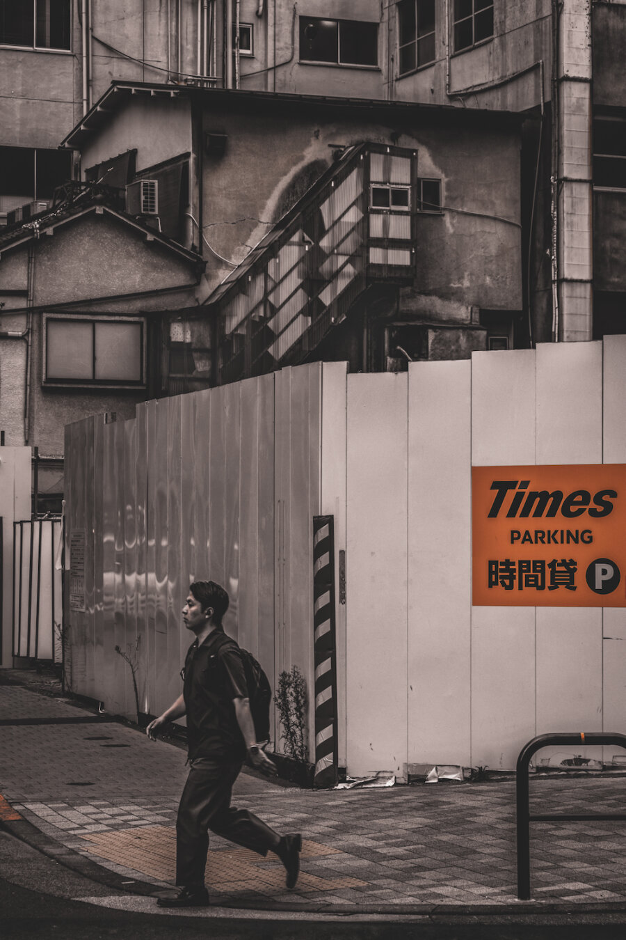 A man walks past a white construction fence with an orange Times Parking sign in Kojimachi, Chiyoda. Behind the barrier, aged buildings and a shadowed outdoor staircase create a quiet, nostalgic urban backdrop, blending daily movement with the stillness of Tokyo’s architectural history.