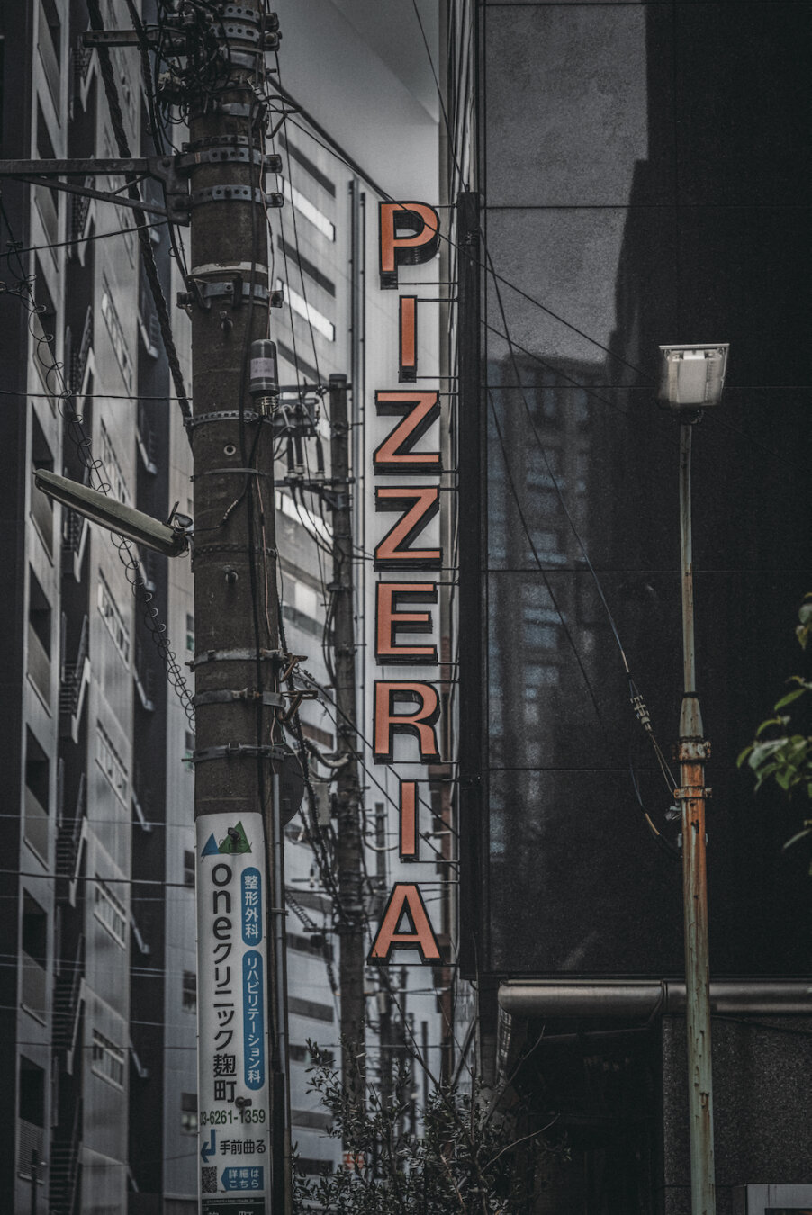 A vertical orange sign spelling PIZZERIA hangs along a sleek dark facade in Kojimachi, Chiyoda. Beside a utility pole wrapped with Japanese advertisements and tangled wires, the sharp typography adds a subtle poetic warmth to the moody, structured streetscape of central Tokyo.