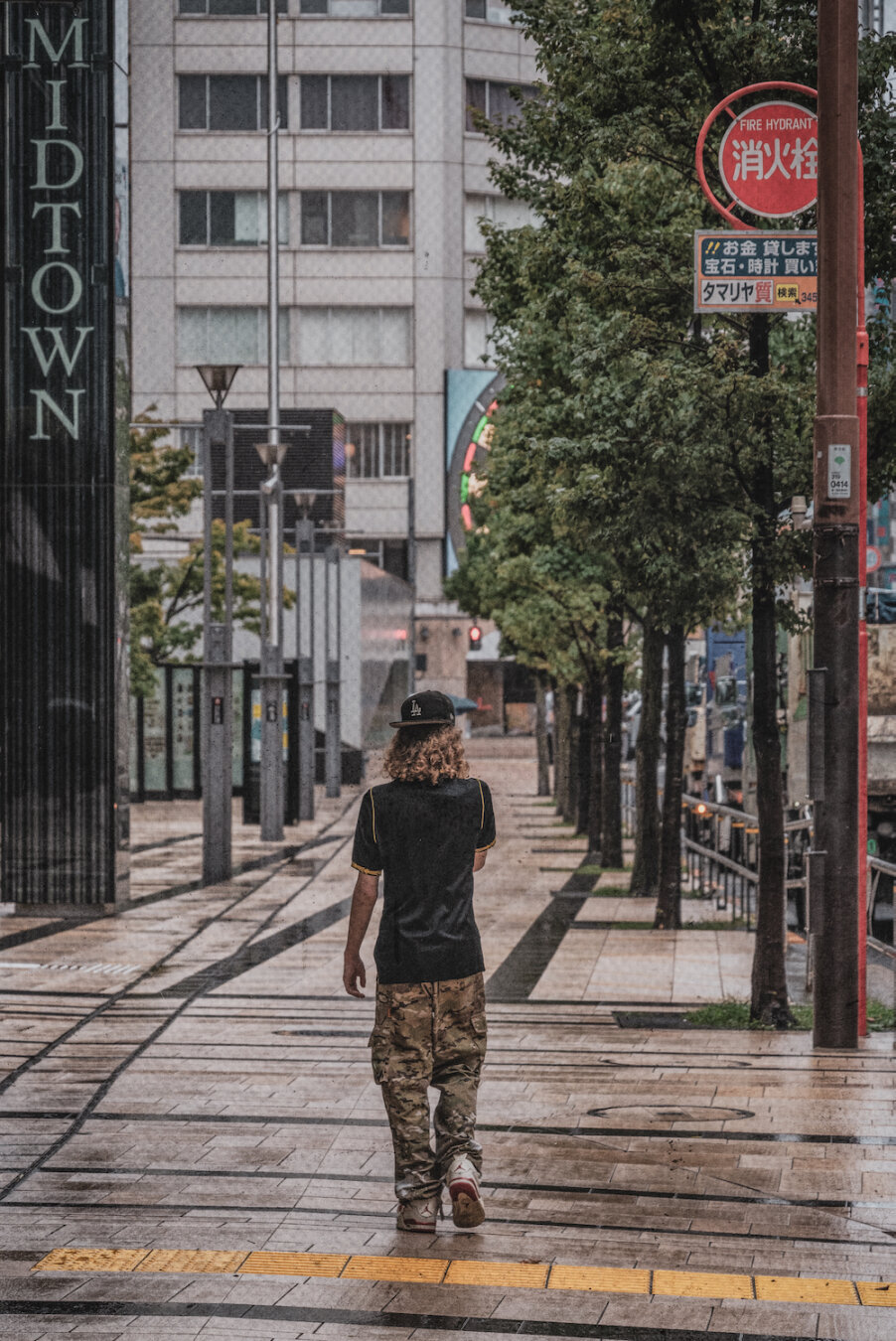 A solitary person with long curly hair wearing a baseball cap, black shirt, and camouflage trousers walks away along a damp pavement near Midtown. Framed by leafy trees, street signs, and modern architecture, the quiet urban scene blends casual street fashion with the calm rhythm of the city.