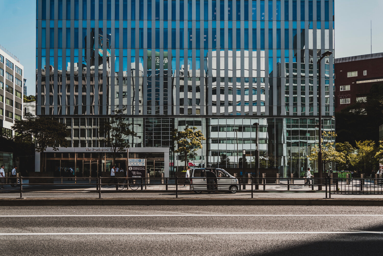 A wide cityscape shot where Nagatacho, Chiyoda Ward, meets Akasaka, Minato Ward. A modern glass facade reflects surrounding buildings, creating a layer of urban scenery above the street level. Cars and pedestrians travel along the road beneath the grand, reflective architecture bathed in clear sunlight.