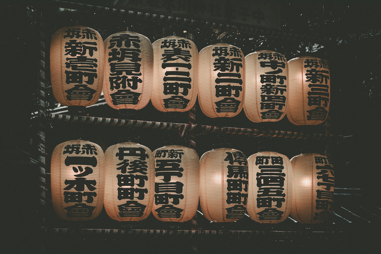 A tranquil morning scene at a shrine, where rows of traditional Japanese paper lanterns, inscribed with local distinct names in red and white kanji, hang glowing softly. Their warm light pierces the remaining darkness, creating a solemn atmosphere in Akasaka, Tokyo.
