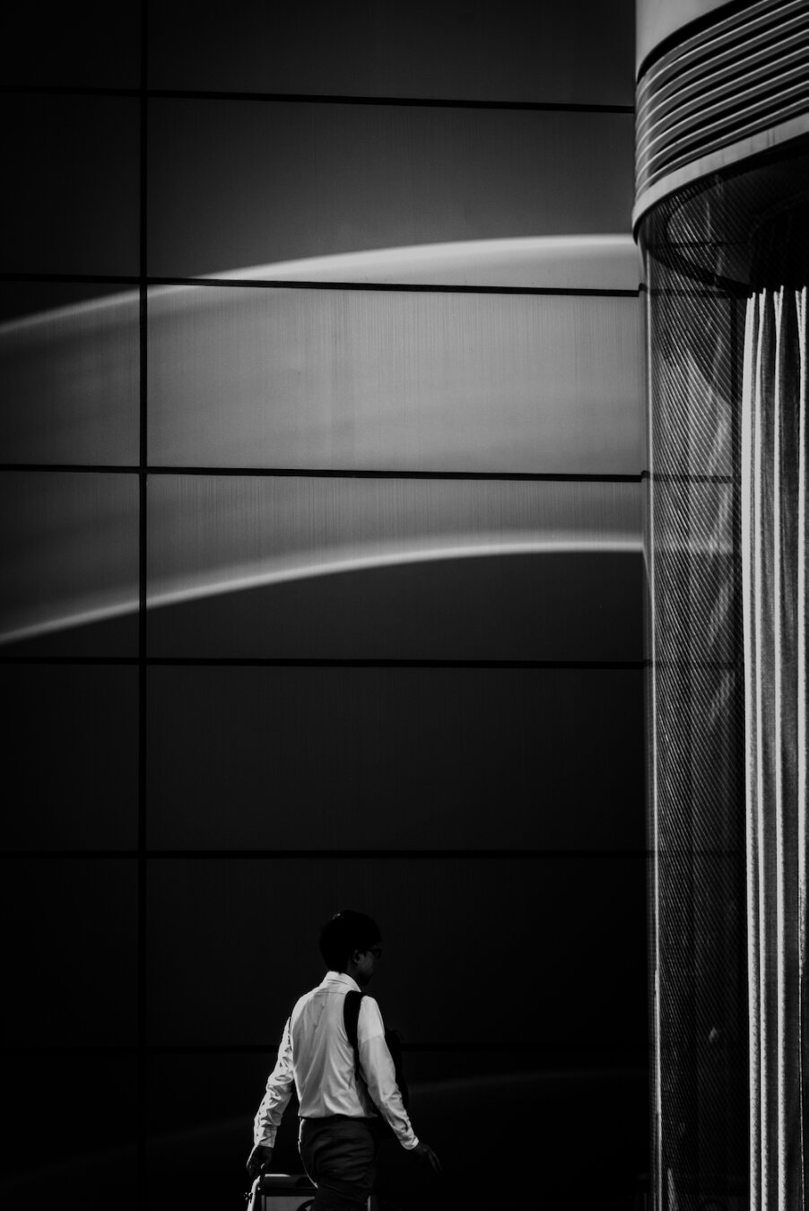 A monochrome vertical shot of modern architecture with sweeping, luminous reflections painted across a glass facade. At the bottom, a solitary figure walks carrying a bag, set against the bold geometric lines and dramatic light, creating an impressionist canvas within an urban landscape.
