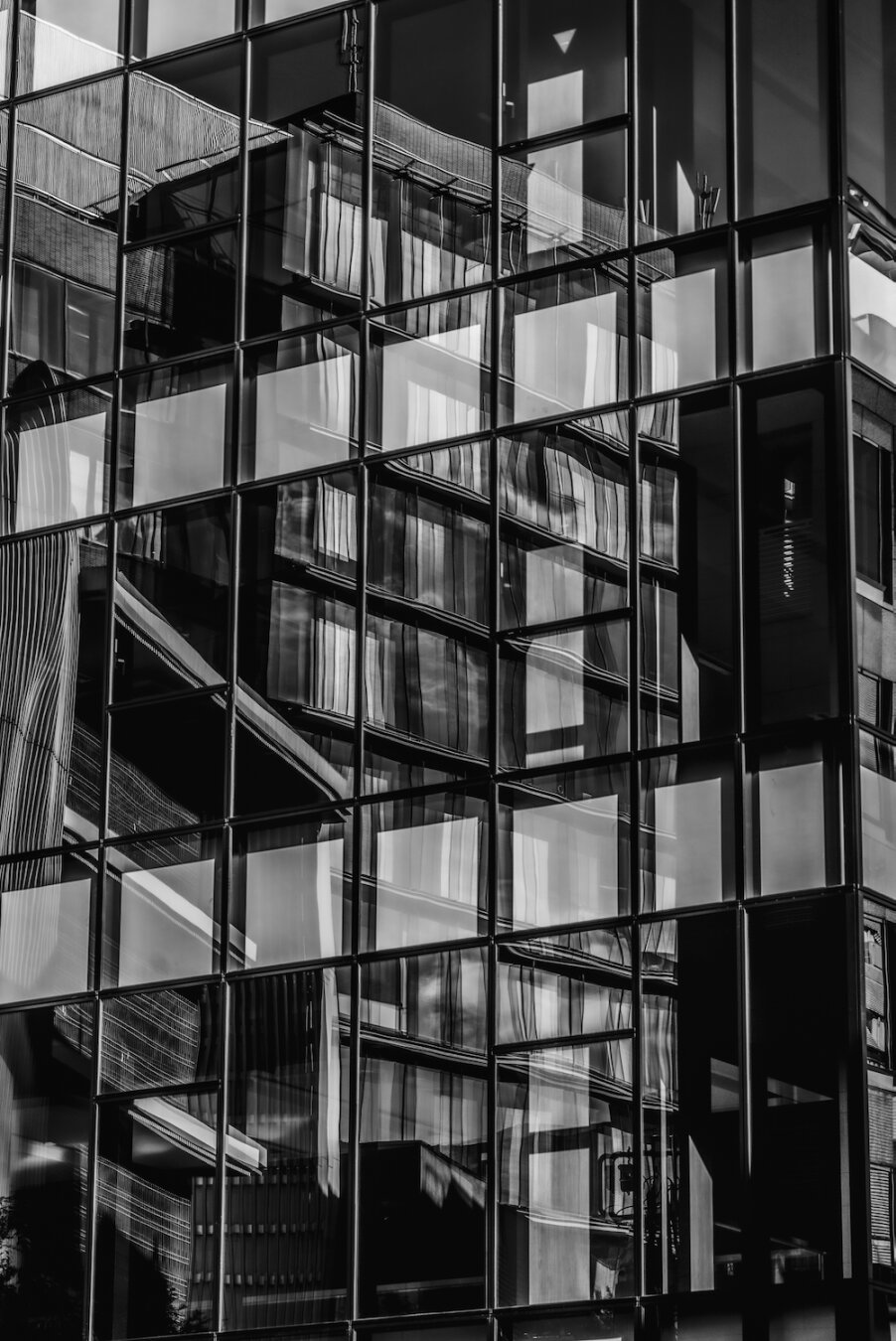 A stark, high-contrast monochrome photograph capturing a geometric facade of a modern glass skyscraper. Sharp sunlight illuminates portions of the grid, creating fragmented reflections and long, dark, distorted forms of surrounding structures on the mirrored panes. The composition is a complex interplay of vertical lines and complex patterns, evoking a fractured, shimmering urban memory.