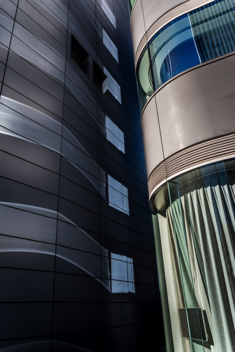 A vertical shot capturing smooth waves of light reflected across the dark panelled facade of a modern skyscraper. Adjacent to the building, a curved glass structure reveals elegant interior drapery. The composition harmonises fluid, sweeping reflections with rigid geometric architecture, transforming urban surfaces into a serene piece of abstract artwork.