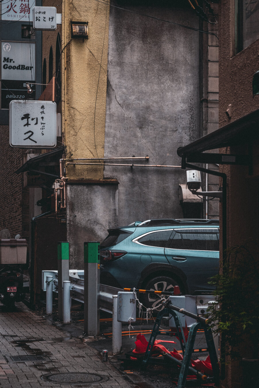 A teal car is parked in a narrow hourly car park tucked neatly between aged city buildings along a slender back alley. Framed by weathered concrete walls, vintage shop signs, and street fixtures, the quiet scene captures a peaceful, hidden pocket of modern urban life resting within the city's dense architecture.