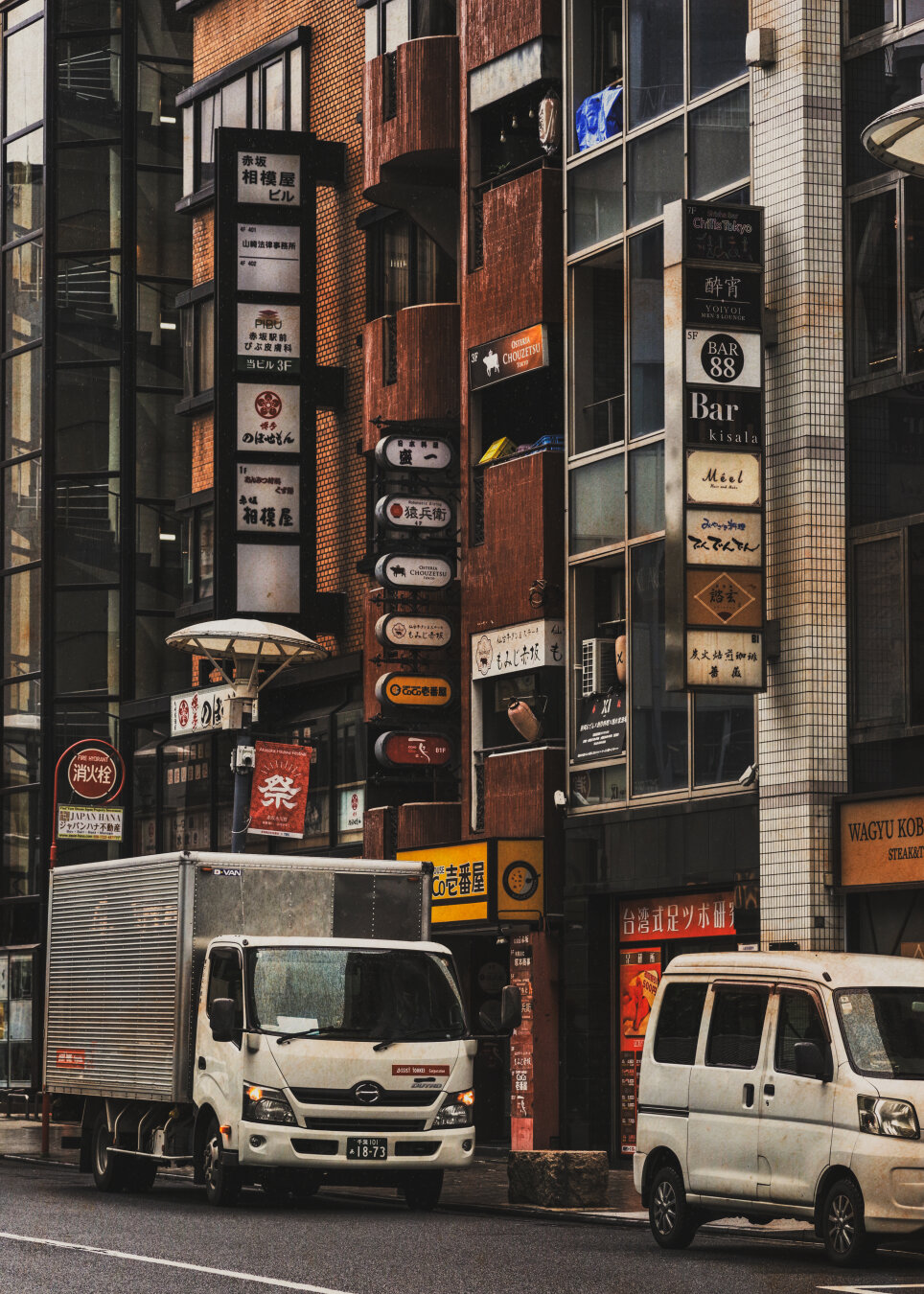 A delivery truck and a white van park along a bustling street in Akasaka, Tokyo, lined with retro brick buildings, shop signs and reddish festive banners, capturing the quiet pulse of urban daily life.