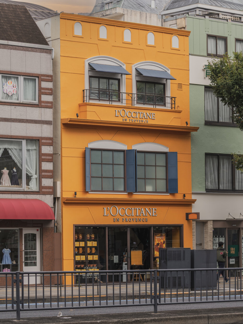 A vibrant yellow three-storey building facade of the L'Occitane shop in Minami-Aoyama 4-chome near Omotesando, Tokyo. The structure features blue window shutters, small awnings, elegant balcony railings, and large display windows at street level, set naturally between adjacent buildings under a soft, overcast city light.