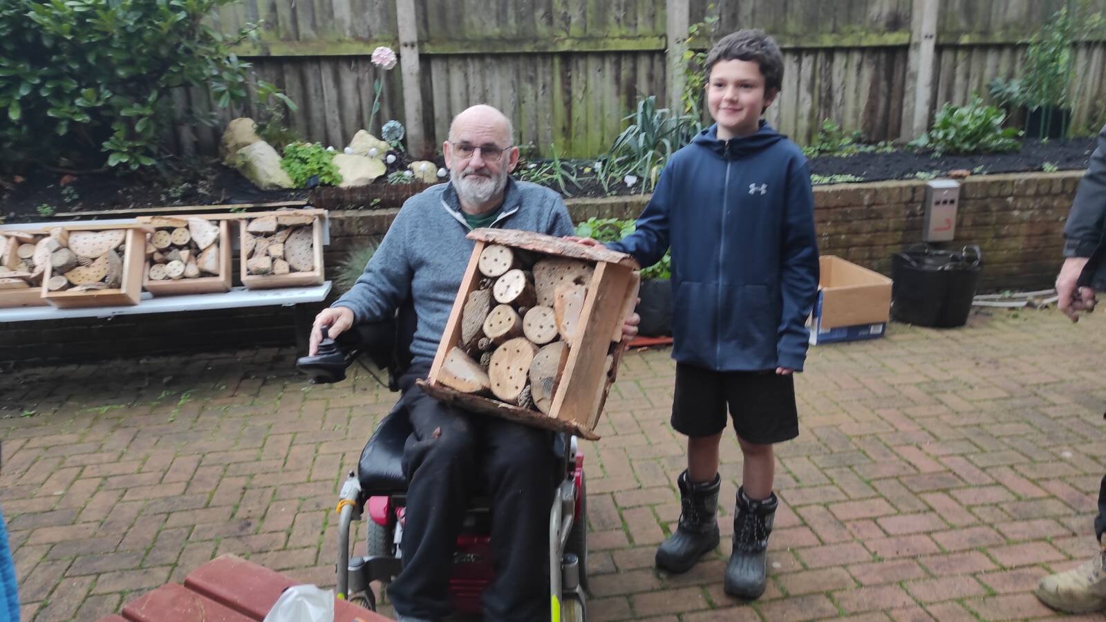 Man in wheelchair holding bug hotel, smiling boy standing next to him