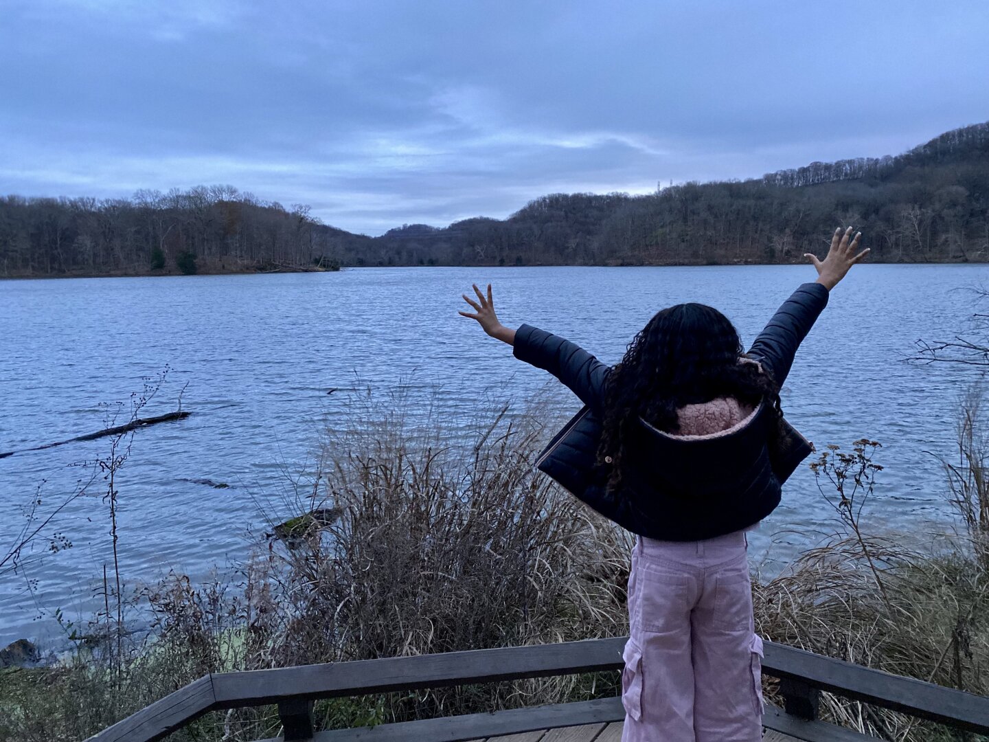 A small very curly haired child wearing pink pants and a puffy black jacket holds her hands up towards the sky in front of a large lake. The sky and the lake are the same color, dark grey blue as the sun descends way down below the hills in the background