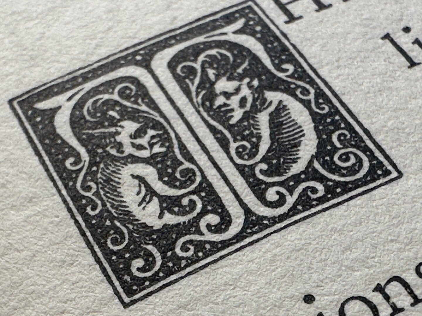 Close up of black stylized letter T with two devil characters starting a sentence on a white page in an old book.