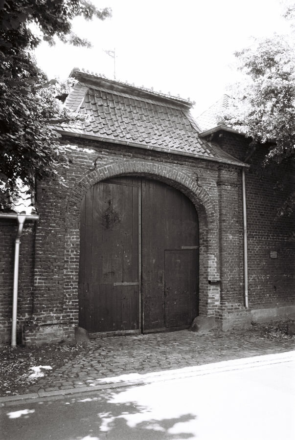 A brick gate with wooden doors, a tile roof, and a small pennant on top. The entryway is paved with large cobblestones.
