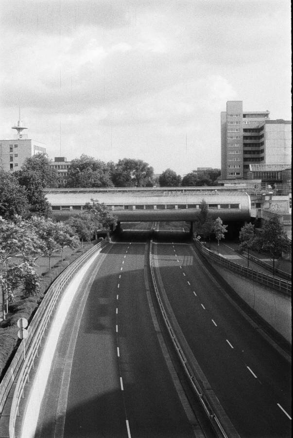 A parkway 4 lanes ride runs under a modernist pedestrian bridge with shops on it. There are no cars on the road.