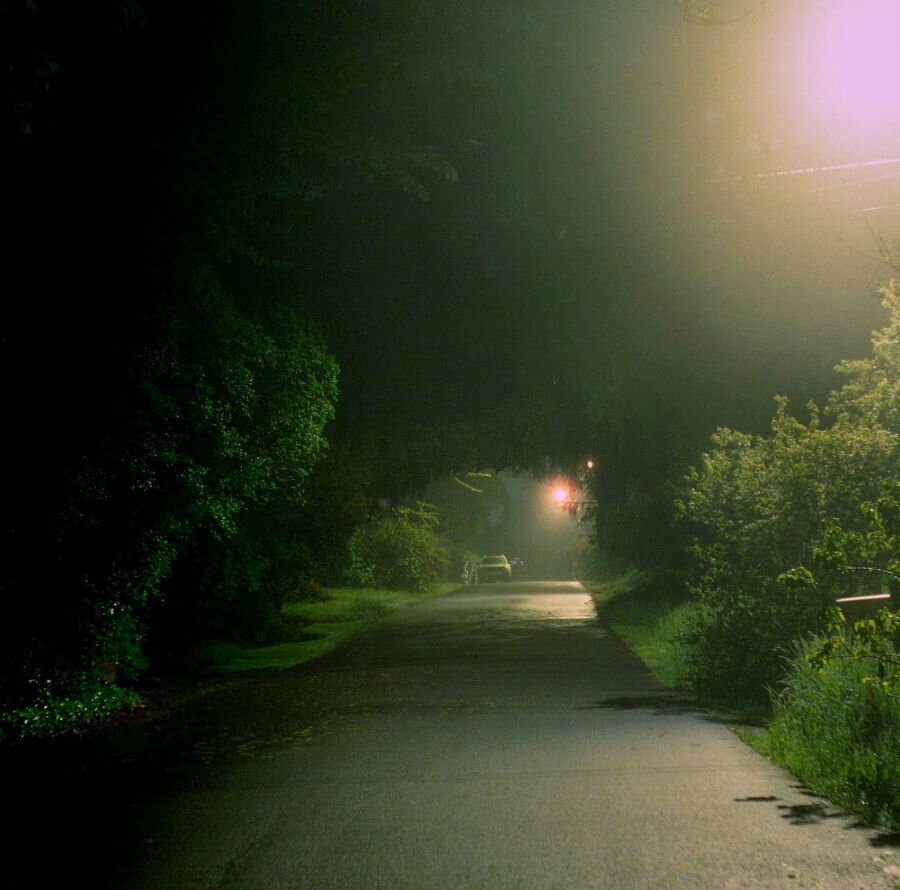 Looking down a mostly-empty alleyway lit by only two streetlamps. There are houses but they aren't visible. There is a lone car parked in the distance.