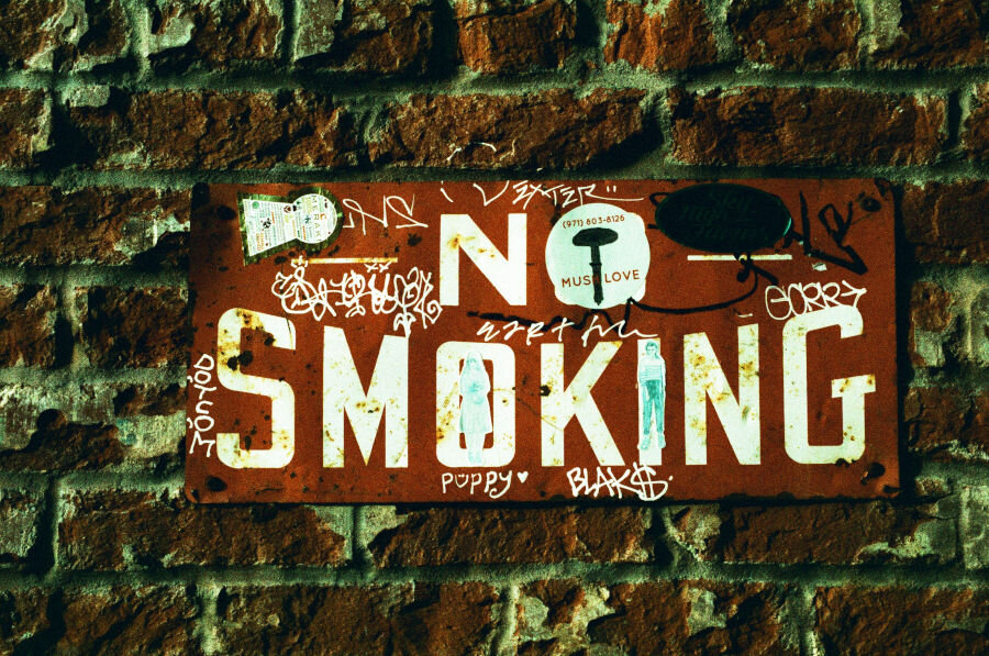 A No Smoking sign on a brick wall. It has been written and drawn on, and had stickers applied.