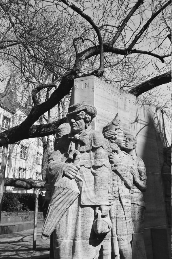 A stone block sculpture under a tree with several figures carved on it in high relief. The central figure is a man with an umbrella in a long coat.