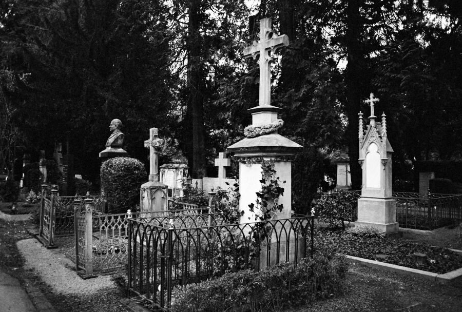 A tall tombstone topped by a cross is surrounded by other monuments, some simple, some Gothic, one with a bust.