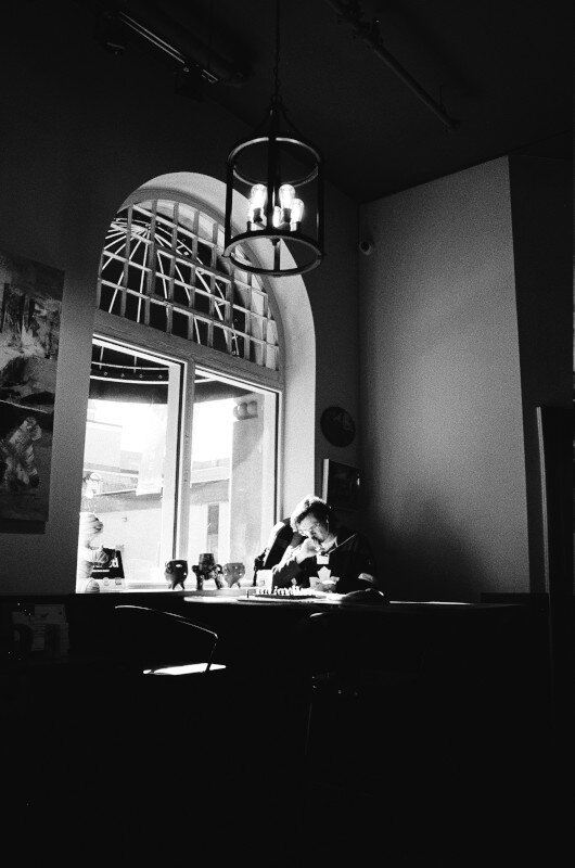 A man sits at a table in a cafe. Beside him is a window with a rounded top. Above him is an iron chandelier. The room is relatively dark.