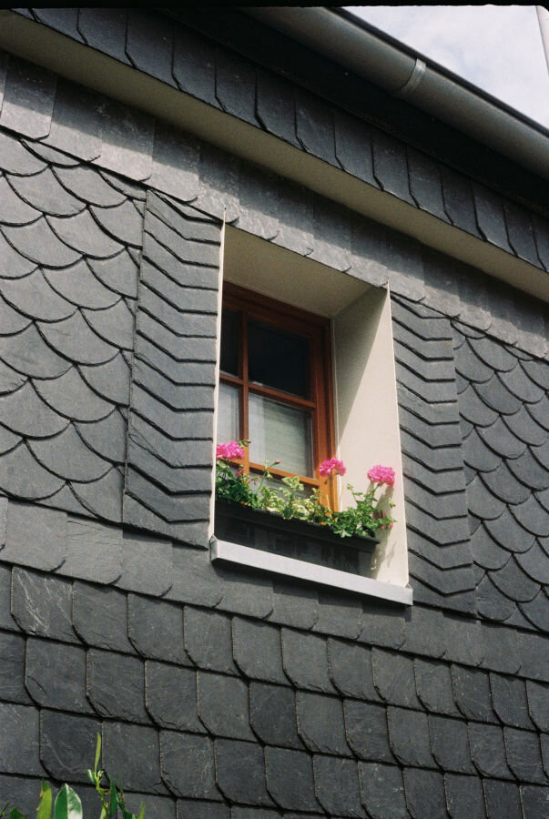 An exterior wall covered in slate. In the window is a flower box full of pink flowers.