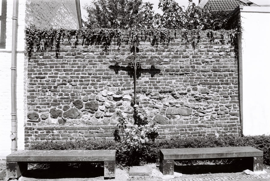 A section of wall with brick filled in by random stones. A wrought iron cross hangs on it and below grows a rose bush. In front of the wall are two benches.