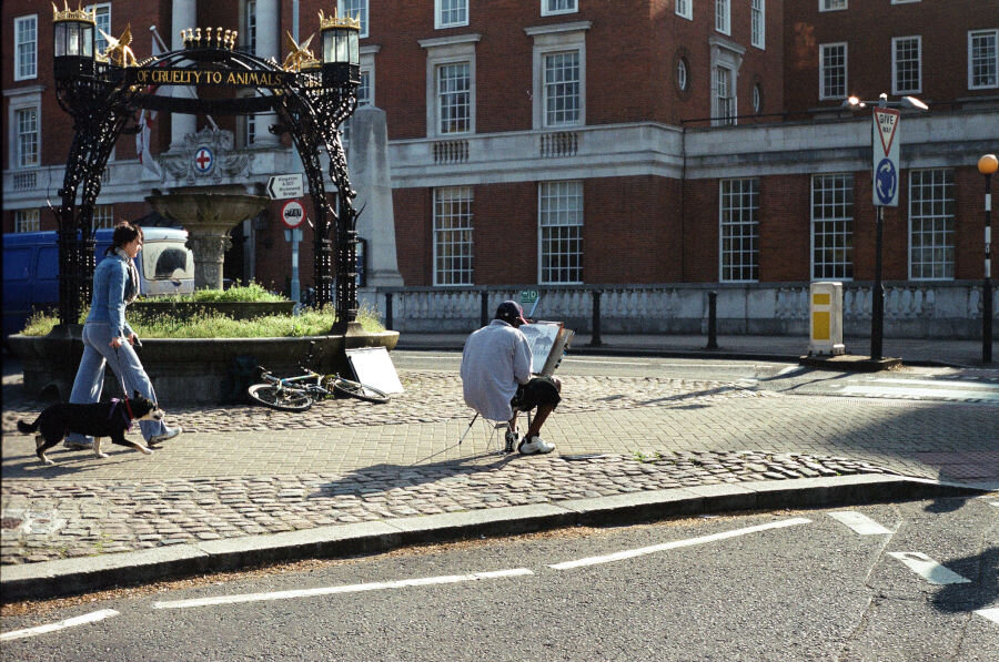 A dark-skinned man in a hat sits in a traffic island with an easel and paper as a woman with a dog walks by.