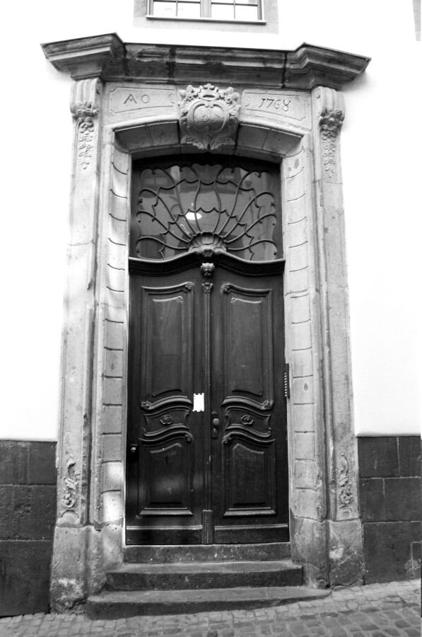 A stone doorway with a crest and the date AD 1768. The door itself has the seashell-like flourishes of the Rococo era full of organic curves with an elegant window above it.
