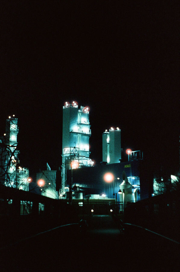 Tall buildings loom in the distance. They seem green-blue in the light. There are marker lights too, that glow with red halation.