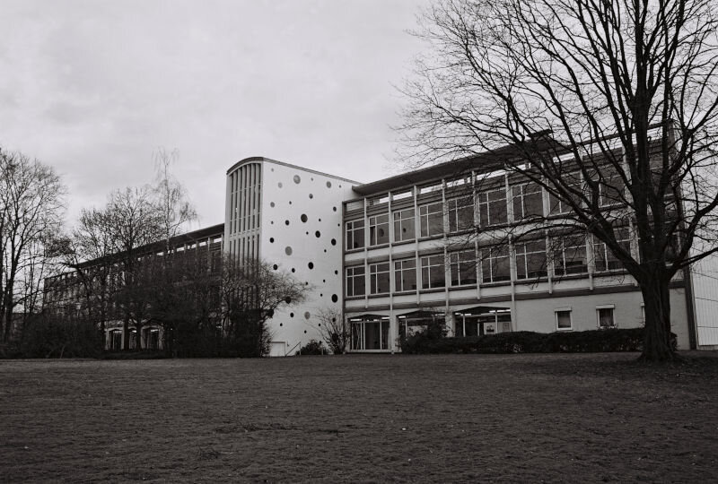 A modern building that is mostly plain boxes but there is a center section with large windows, a curved roof, and smaller round windows at varying sizes and intervals on the side.