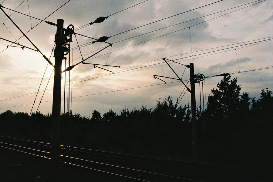 Overhead wires and bushes in silhouette with sun glinting off railroad tracks. Above is a cloudy sky with sun coming through.