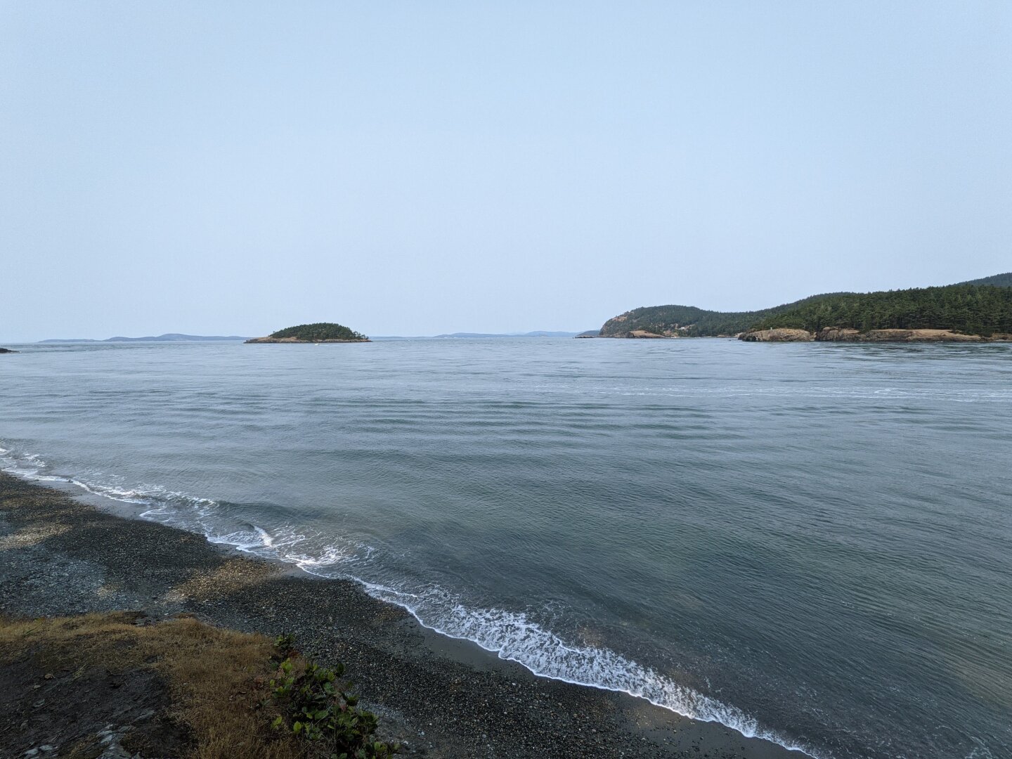 A view of a rocky beach and ocean with a small island and a forested landmass in the distance under a pale sky