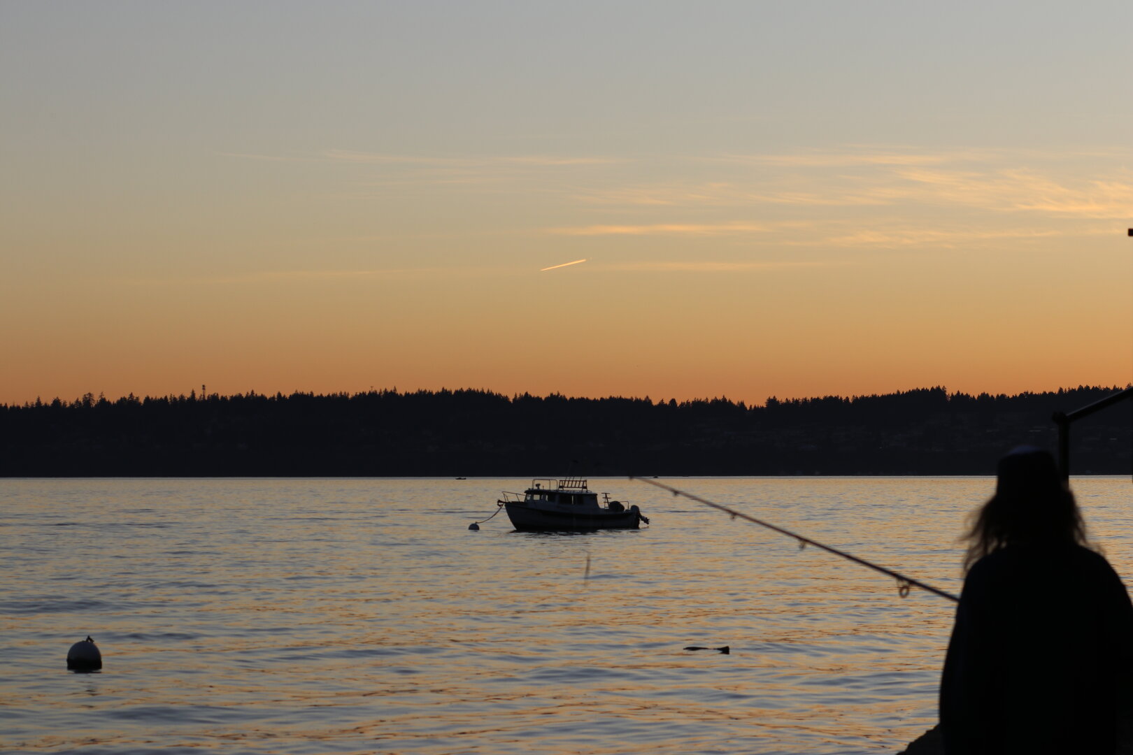 A silhouette of a person fishing from a shore or dock, with a small boat anchored in the water in the background, set against a warm orange and yellow sunset sky over a dark, tree-lined horizon