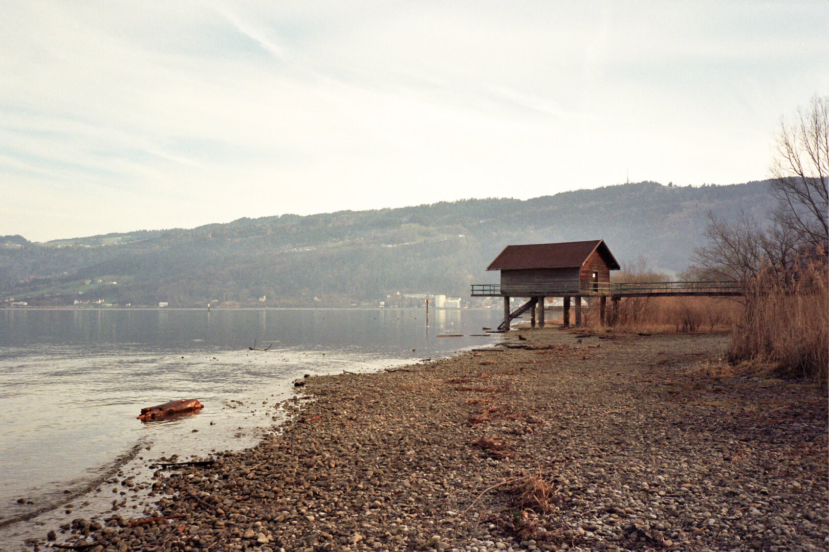 Wooden pier with small wooden house on stilts at the shore of Lake Constance in Bregenz, pebble beach and wooded hills in the background. *** Holzsteg mit kleinem Holzhaus auf Stelzen am Ufer des Bodensees in Bregenz, Kiesstrand und bewaldete Hügel im Hintergrund.