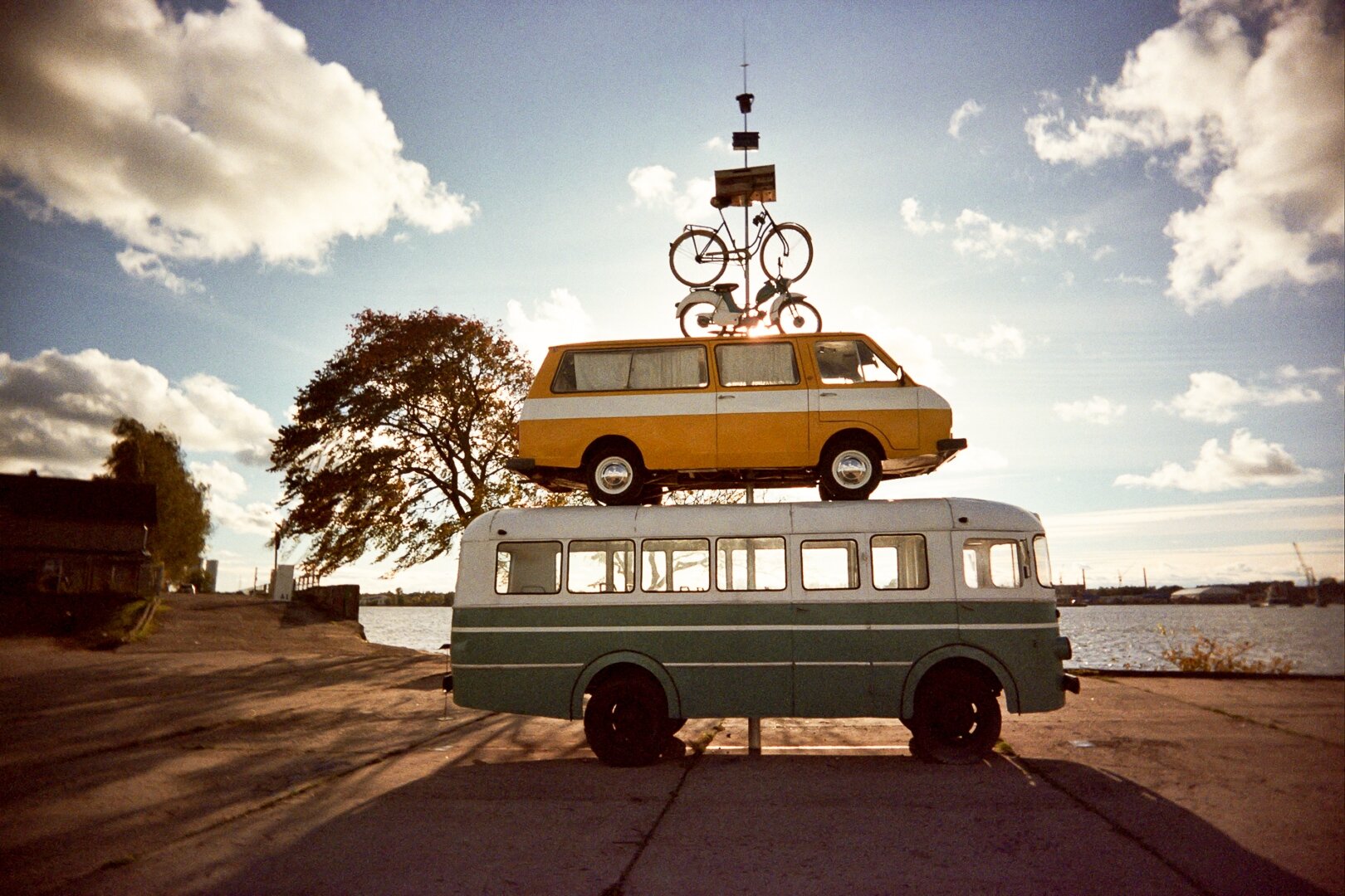 A green bus with a yellow-and-white minibus on its roof, topped by a bicycle, under a cloudy sky in Riga. *** Ein grüner Bus mit einem gelb-weißen Kleinbus auf dem Dach, auf dem ein Fahrrad montiert ist, vor bewölktem Himmel in Riga.