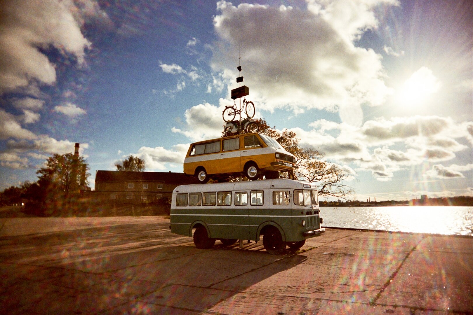 A green bus with a yellow-and-white minibus on its roof, topped by a bicycle, under a cloudy sky in Riga. *** Ein grüner Bus mit einem gelb-weißen Kleinbus auf dem Dach, auf dem ein Fahrrad montiert ist, vor bewölktem Himmel in Riga.