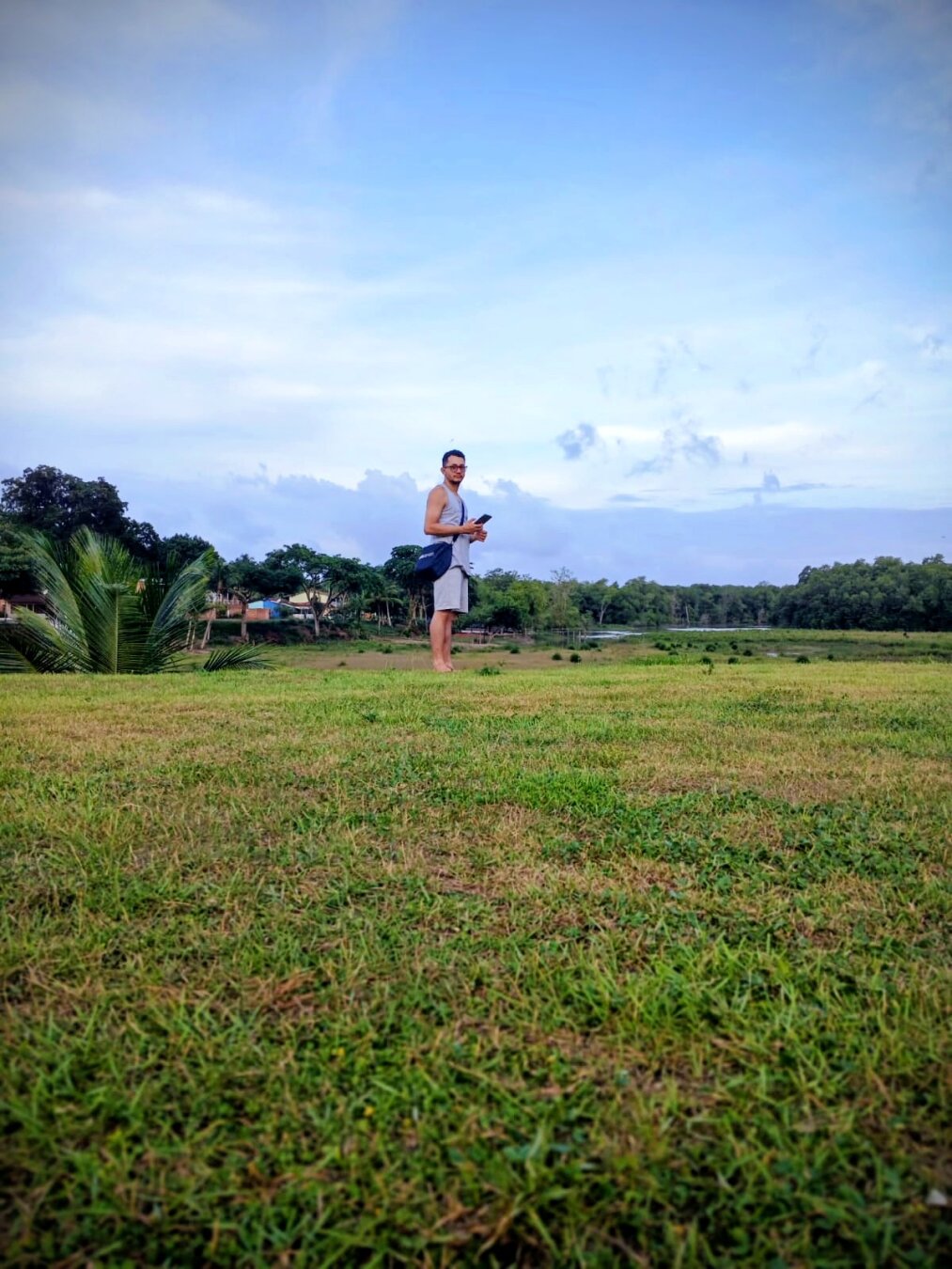 Nesta fotografia, estou posicionado centralmente, a uma distância considerável da câmera, o que me faz parecer diminuto diante da vasta paisagem natural que me cerca. Ao fundo, é possível observar a vegetação de manguezal, enquanto eu estou sobre a grama da encosta do leito da maré, que se encontra seca no momento. É uma fotografia pessoal e ampla, comigo no centro da composição.