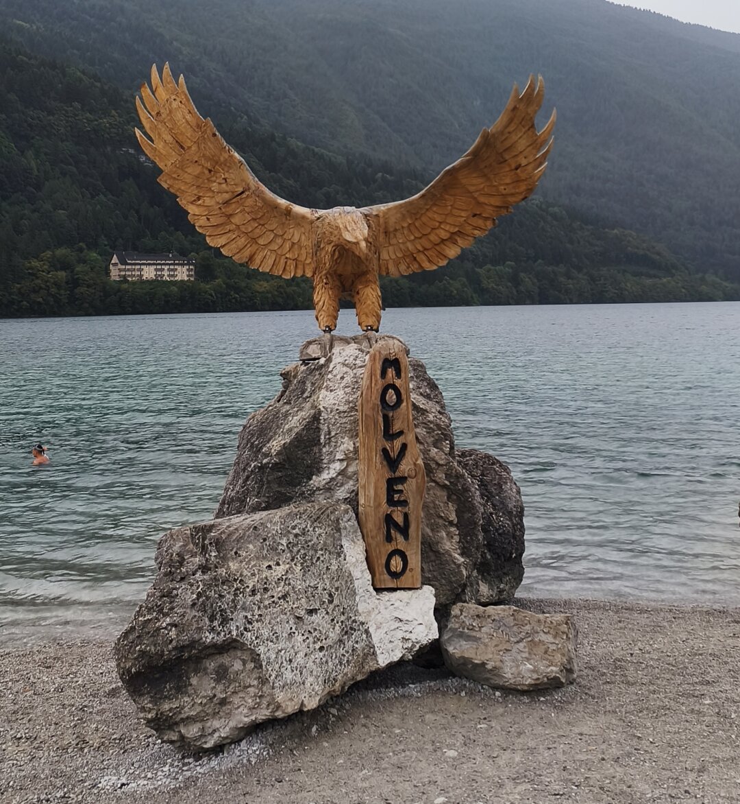 Die Holzskulptur eines Adlers mit ausgebreiteten Schwingen, am Seeufer, darunter auf einer Holztafeln der Schriftzug "Molveno".