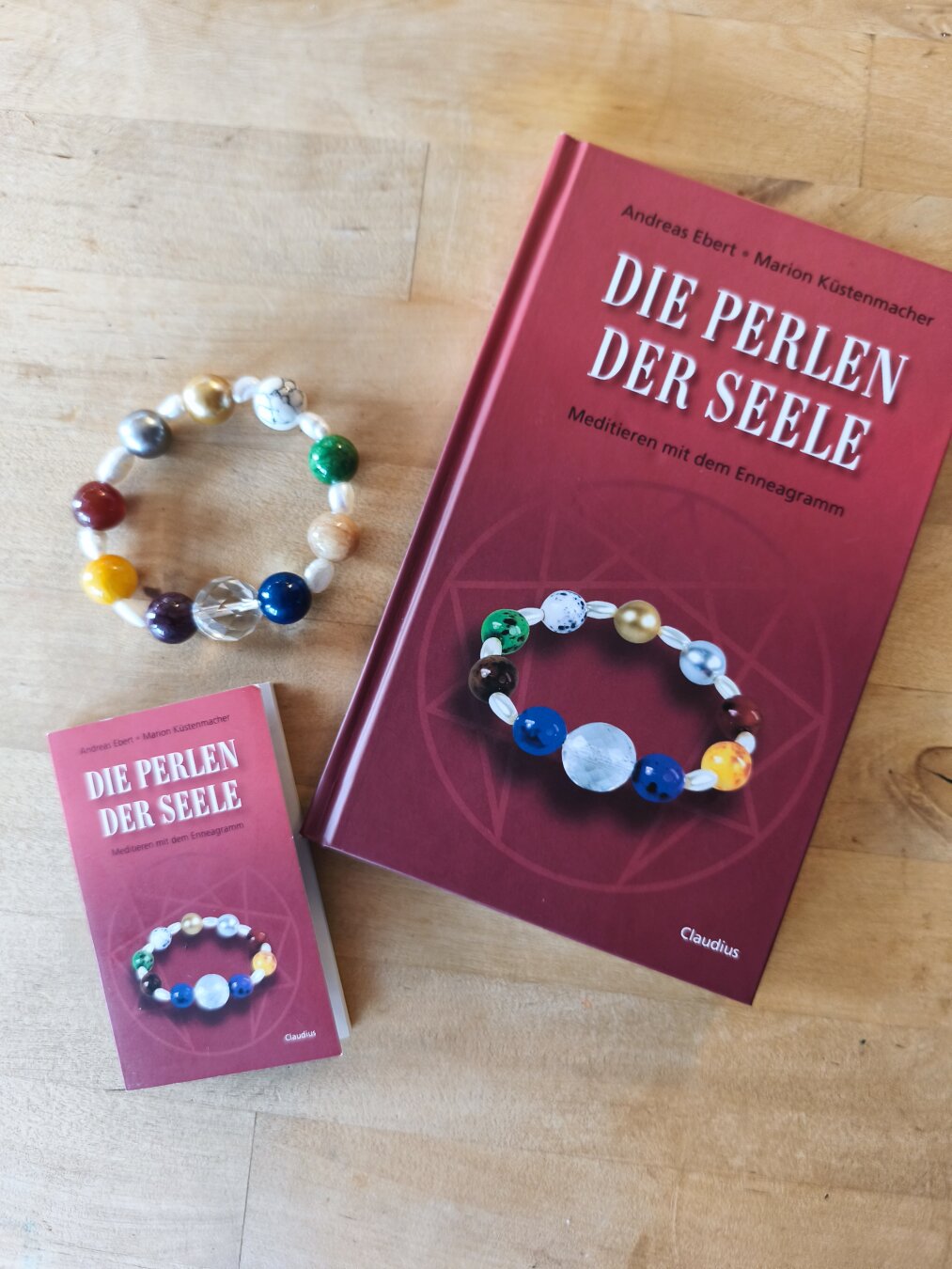 Ein kleines Buch, Titel "Die Perlen der Seele", Autoren Andreas Ebert und Marion Küstenmacher, daneben liegen ein Armband mit verschiedenfarbigen Glasperlen (eine für jeden Enneagrammtyp) und ein Leporello passend zum Buch.