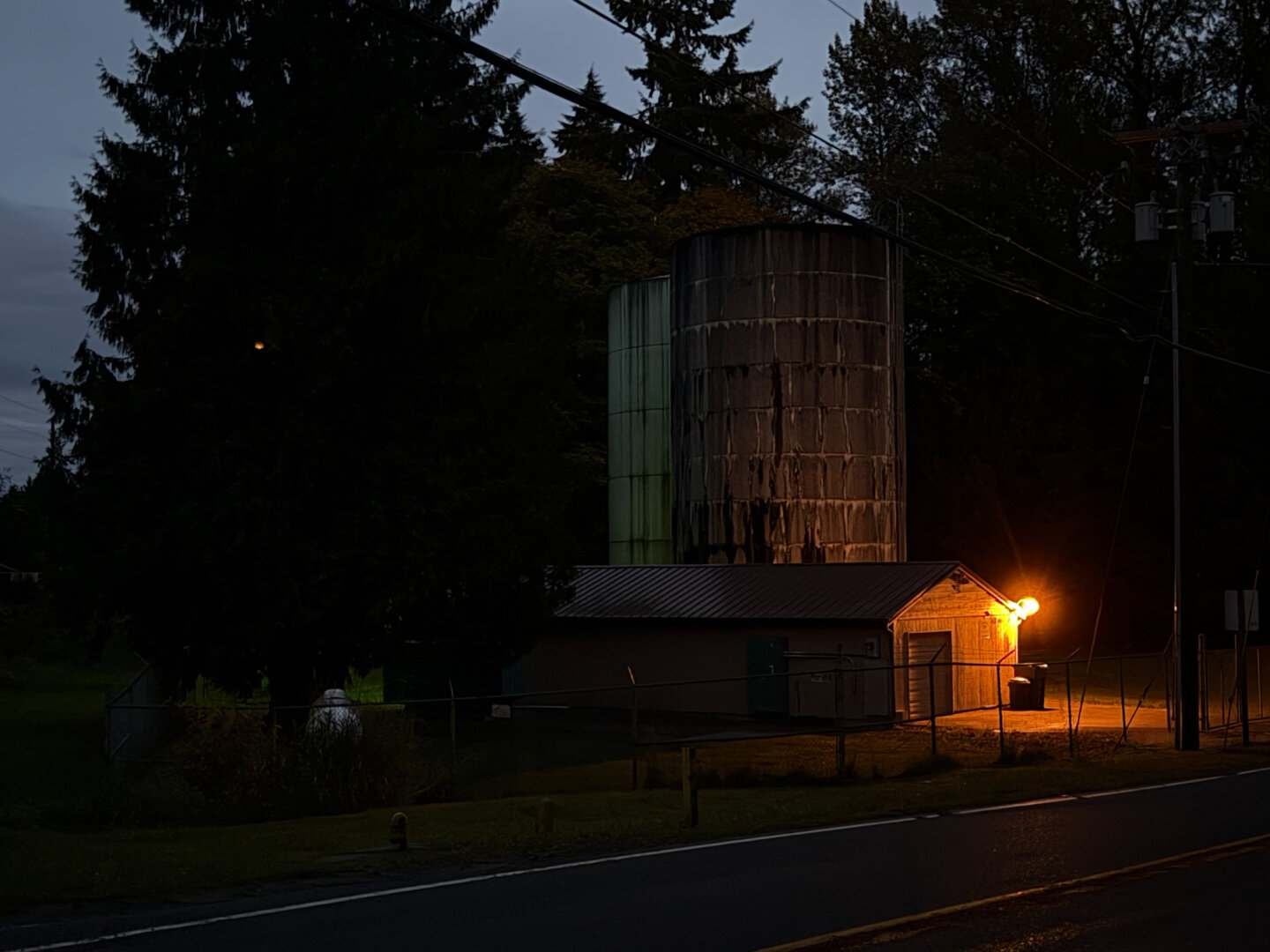 A small service building at night, lit by a single, warm light on its front. Backed by a small silo and coniferous trees.