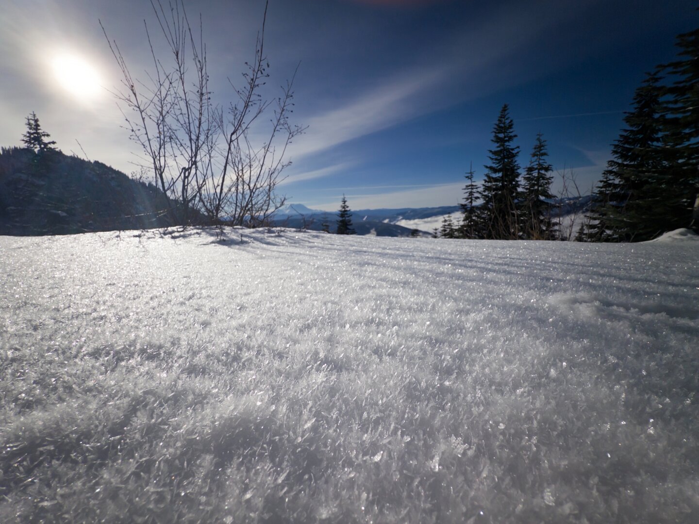 Macro shot of snow, covered in small crystals, in the foreground with some trees and distant mountains in the background. Sun is behind hazy clouds in the corner.