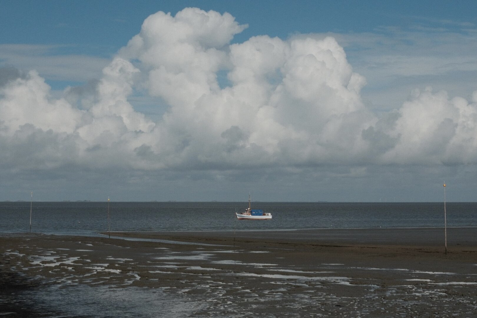 Im Vordergrund ist das dunkle, nasse Watt bei Ebbe zu sehen, durchzogen von Wasserläufen. Am Horizont beginnt das offene Meer. Auf dem Wasser liegt ein kleines, weißes Fischerboot mit einem blauen Aufbau. Es ist festgemacht oder liegt im Schlick. Im Watt stecken mehrere dünne Stangen, die als Markierungen dienen. Der Himmel ist hellblau und zu großen Teilen mit massiven, weißen, bauschigen Cumuluswolken bedeckt, die sich über dem Meer auftürmen.

The foreground shows the dark, wet mudflat at low tide, crisscrossed by water channels. The open sea begins at the horizon. A small, white fishing boat with a blue cabin is situated on the water, either moored or settled in the silt. Several thin poles, serving as markers, are stuck into the mudflat. The sky is bright blue and largely covered by massive, white, puffy cumulus clouds towering over the sea.