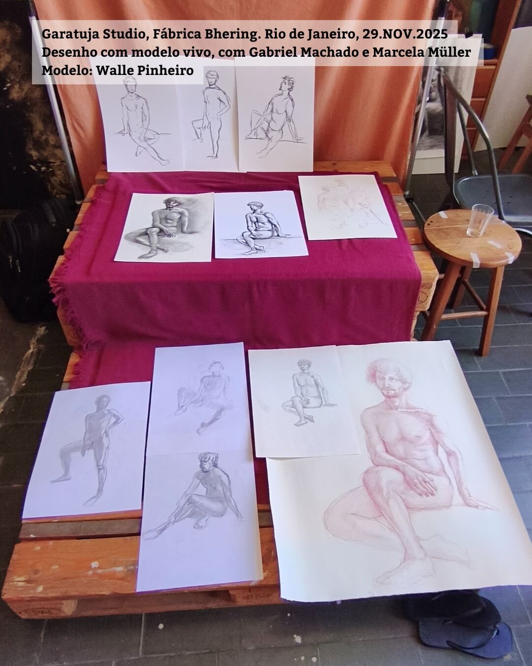 Foto mostra os vários desenhos dos participantes resultantes da sessão de modelo vivo. Os desenhos em papel branco estão cima de um pequeno tablado. Há desenhos a carvão, lápis e um grande com sanguina. O modelo é um homem branco e aparece sentado sobre o tablado na maioria das poses.