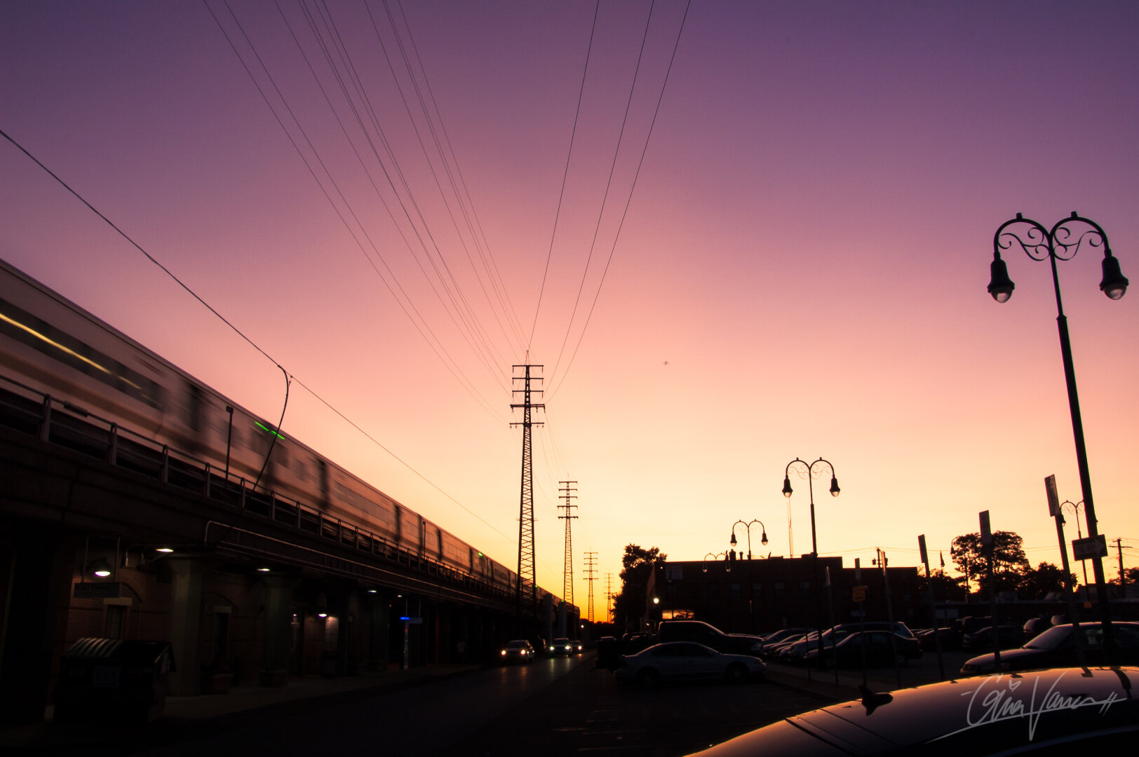 A train is moving across the elevated tracks to the side of the photo, while the rest of the photo is taken up by the purple, pink, and yellow hues of a sunset. These colors are framed by the parking lot and the surrounding structures.