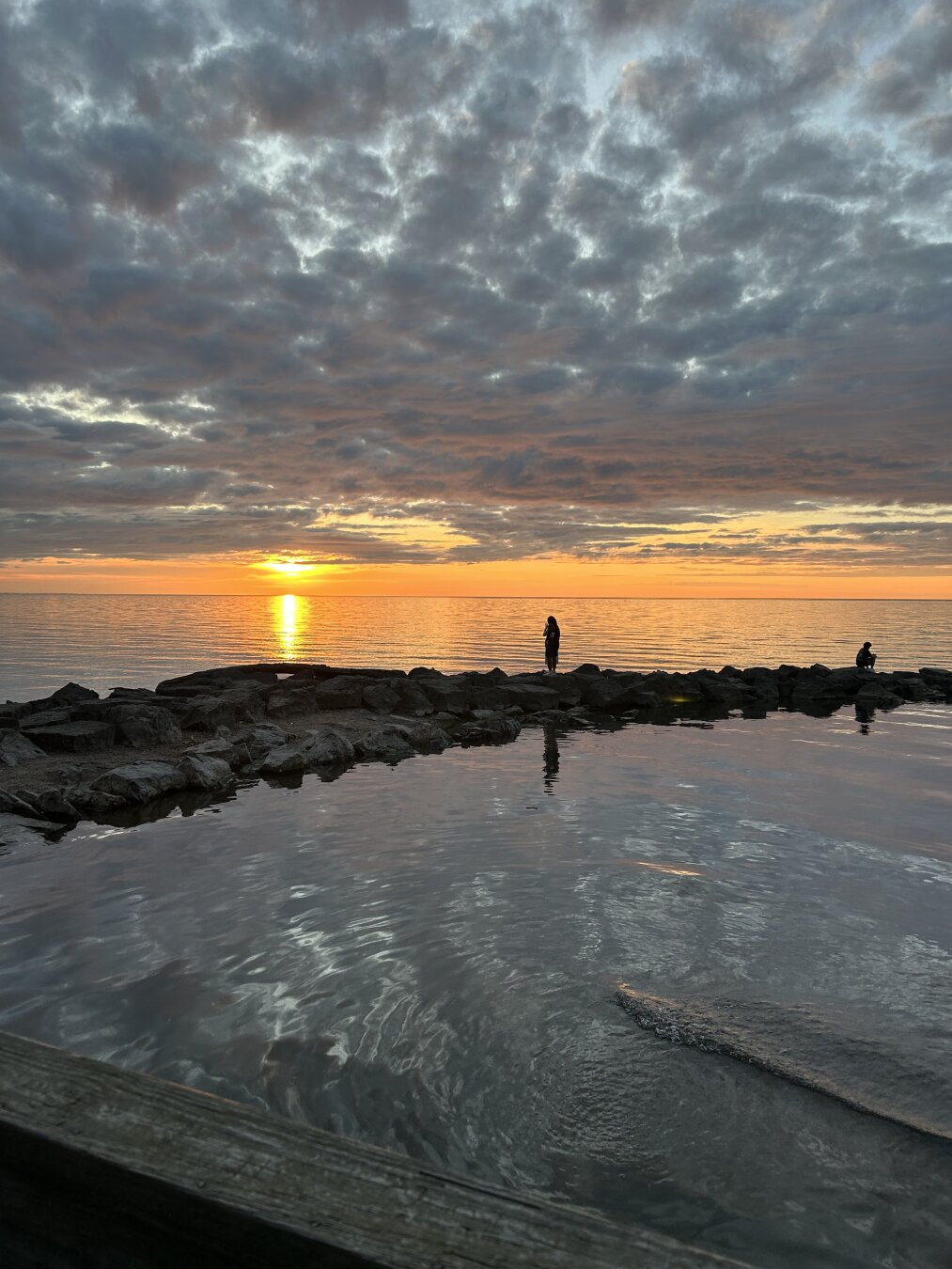 A photo of a summer sunset over Mill Creek and Lake Ontario. Clouds dot the sky as the sun peeks through. There are people standing on the rocky shore, looking out in the distance.