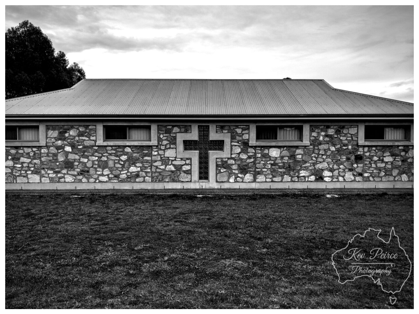 A stark, symmetrical black and white photograph of the side hall or auxiliary building of Manaranga Church.  The structure features a low pitched roof of corrugated iron and a rugged exterior wall constructed from varied, irregular fieldstones.  Centred on the wall is a large, recessed cross made of glass block or heavily framed stained glass, flanked by two horizontal windows on either side. A grassy foreground sits beneath a cloudy sky.