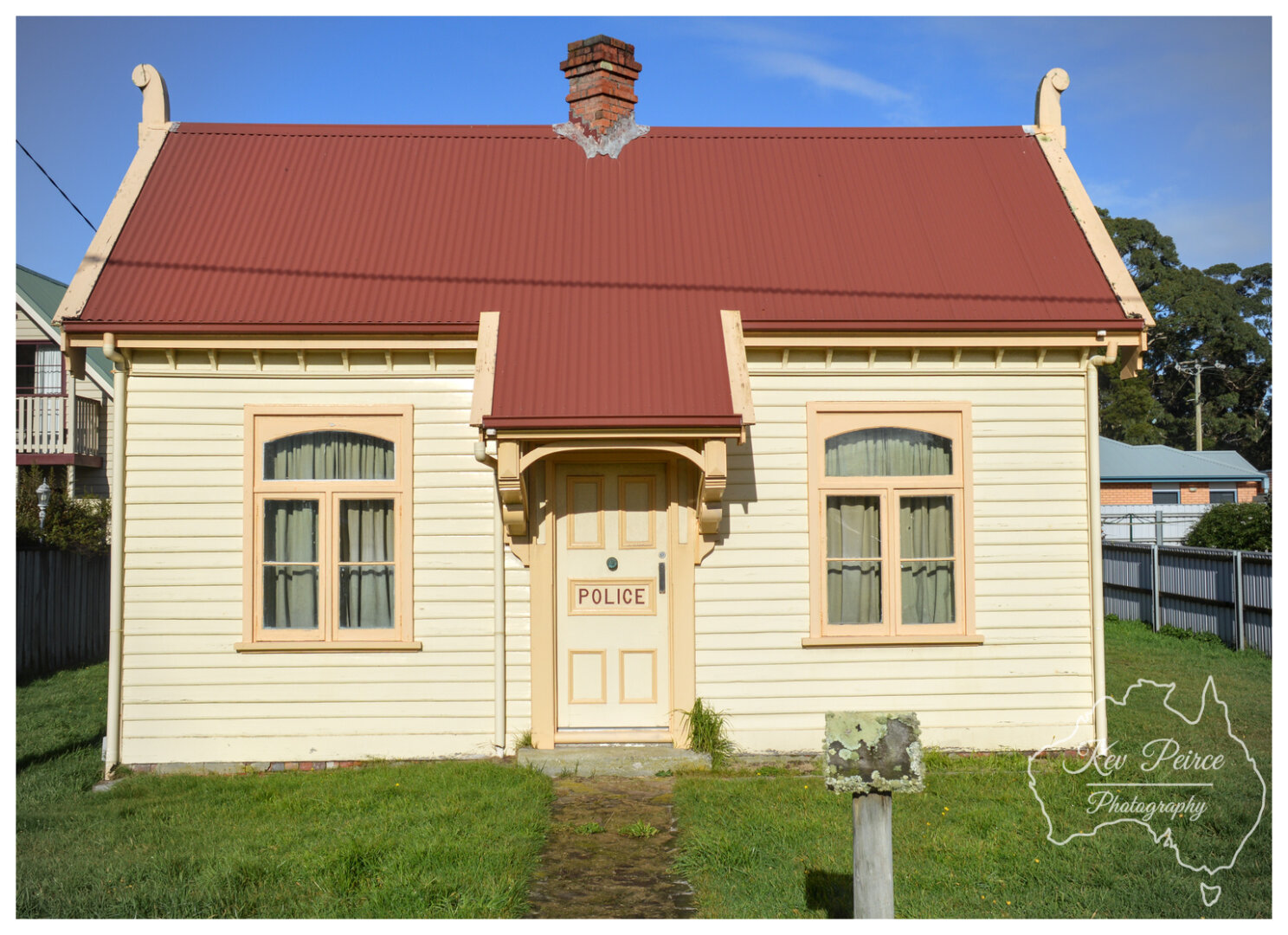 A centred shot of a historic, single story wooden building with light yellow horizontal siding and a reddish brown corrugated iron roof. The door is labelled "POLICE." Two arched windows flank the central entrance.