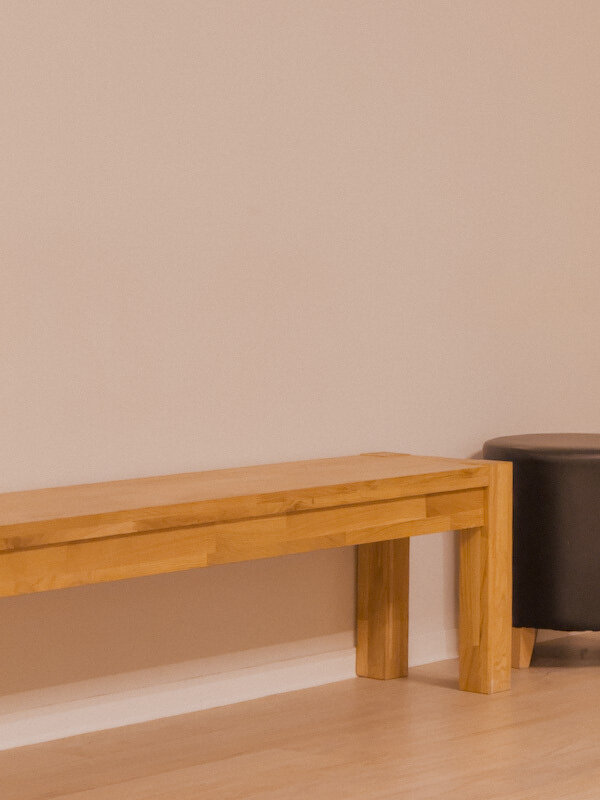 Eine hölzerne Sitzbank steht neben einem runden, schwarzen Hocker auf einem Holzboden vor einer cremefarbenen Wand.