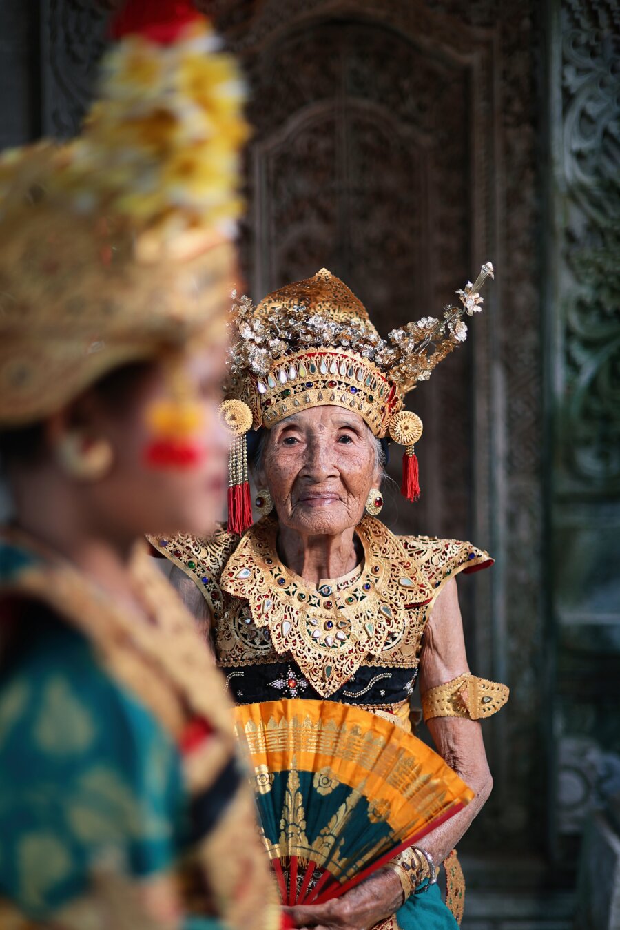 Portrait of the world's oldest traditional Balinese dancer at 106, Niang Soli in traditional attire focused with a younger dancer in the foreground.