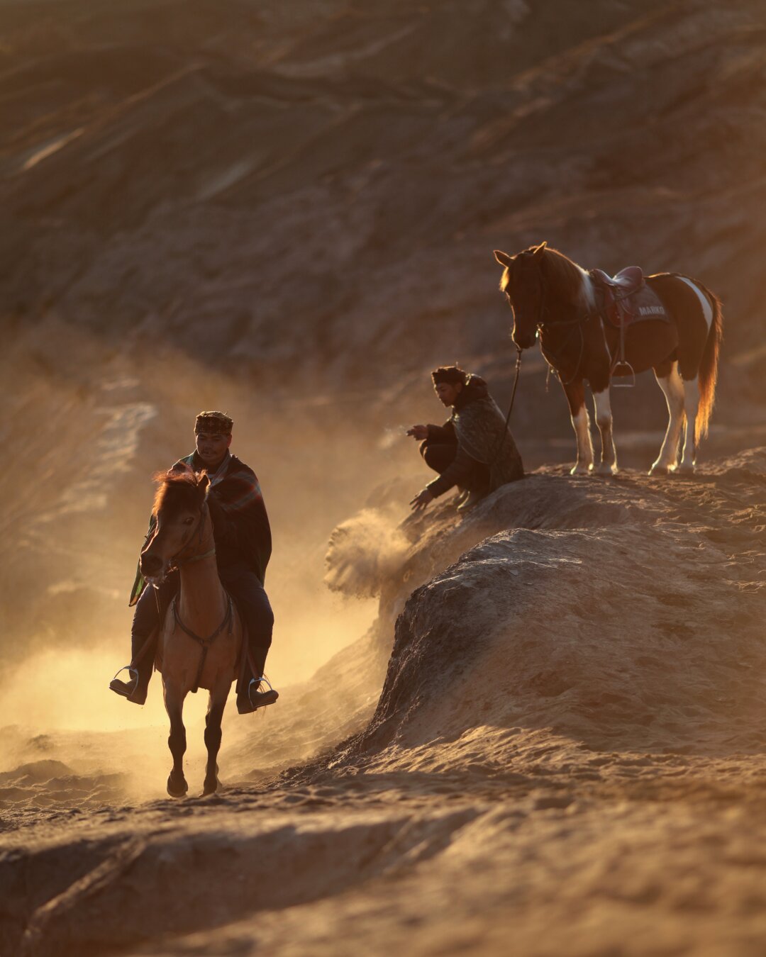 First horseman now trotting towards the camera while the squatting one drops some sand down in the background