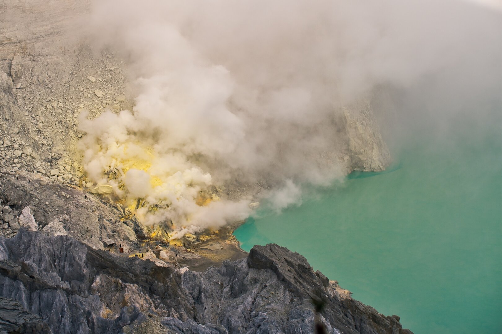 Bright yellow sulphur fumes rise from side of a cyan lake, located inside a volcanic crater