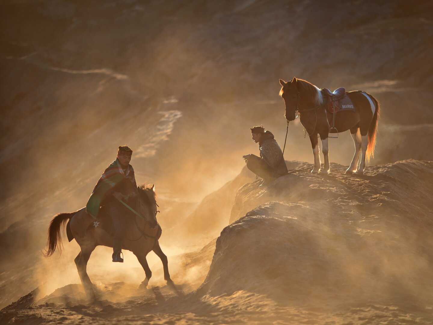 A horseman turns on his horse, while another squats down besides his on a sun-lit sandy volcanic landscape
